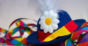 Blauer Hut mit Gänseblümchen und Feder liegt auf bunt gemusterten Luftschlangen und Konfetti